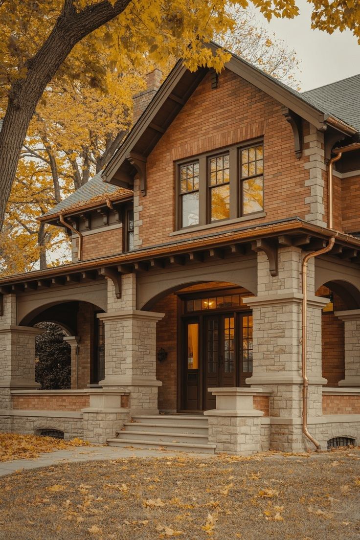 A brick house with a deep eave, stone porch columns, and lit windows, under autumn trees.